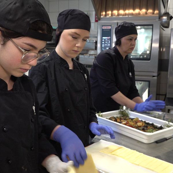 Tre personer i köksuniform förbereder mat i ett skolkök.