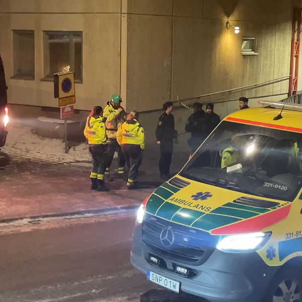 Räddningstjänst och ambulanspersonal arbetar i ett vinterlandskap vid en byggnad. En ambulans står parkerad på gatan, medan flera personer i skyddskläder diskuterar vid en svart minibus. Det är mörkt, och snö ligger på marken.