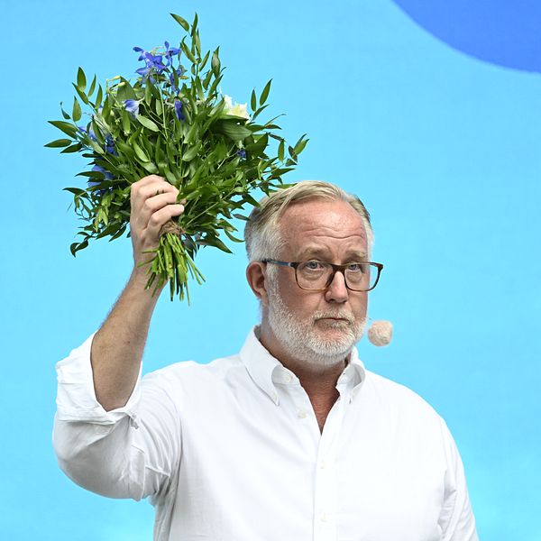Johan Pehrson i vit skjorta håller upp en bukett blommor mot blå bakgrund på scen.