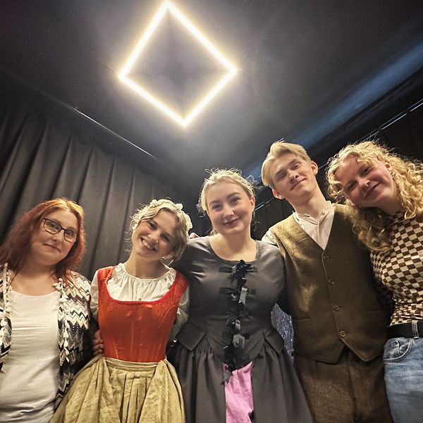 Fem personer i kostym under en ljuskub på scenen, glatt poserande inför en teateruppsättning.