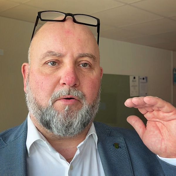 Man i blå kavaj står i ett kontor och håller ena handen upp, glasögon på huvudet.