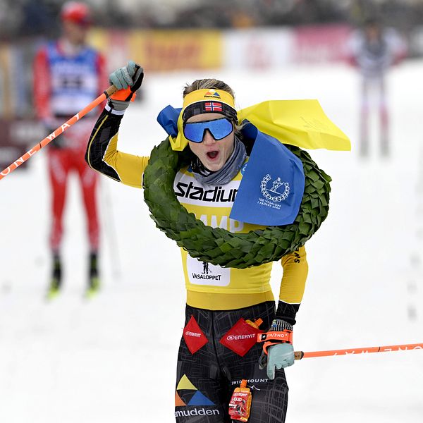 En skidåkare i en gul tävlingsdräkt korsar mållinjen med en laurbärkrans och en gul och blå flagga runt halsen. I bakgrunden syns flera andra skidåkare på en snöig bana.