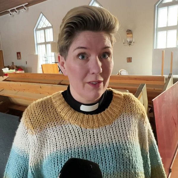 En person iklädd en stickad tröja och en prästkrage står i en kyrka med träbänkar och ljusa fönster i bakgrunden.