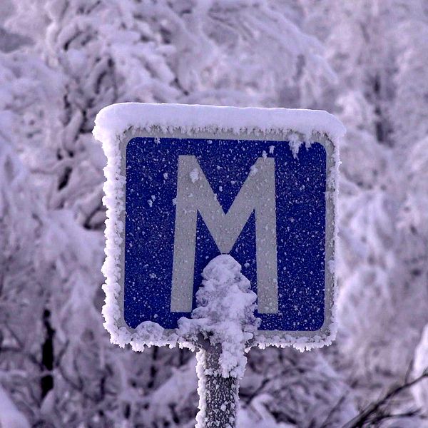 en frostig vägskylt som det står M på i ett vintrigt landskap med snö på träden