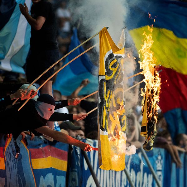 Fotbollsfans bränner motståndarens flagga under match. Flera personer bär munskydd.
