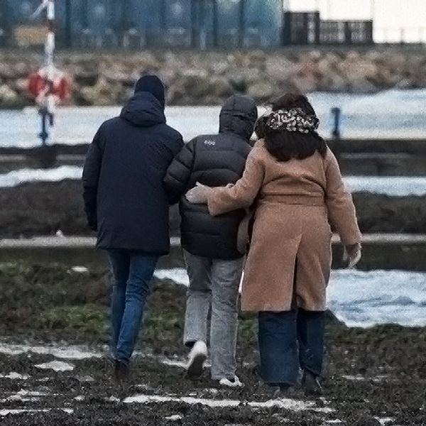 Tre personer promenerar längs en kustlinje vid havet, insvepta i vinterkläder. I bakgrunden syns en byggnad nära vattnet och vågor slår mot stranden.