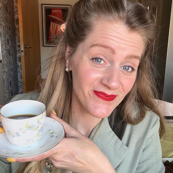 Kvinna i 1940-talsklänning och rött läppstift håller en kopp kaffe i ett kök med blommor och bakverk på bordet.
