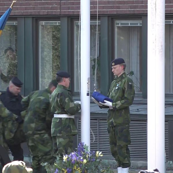 Två militärer hissar Nato-flagga, bakom dem kommer fem personer bärande på man i militärkläder.