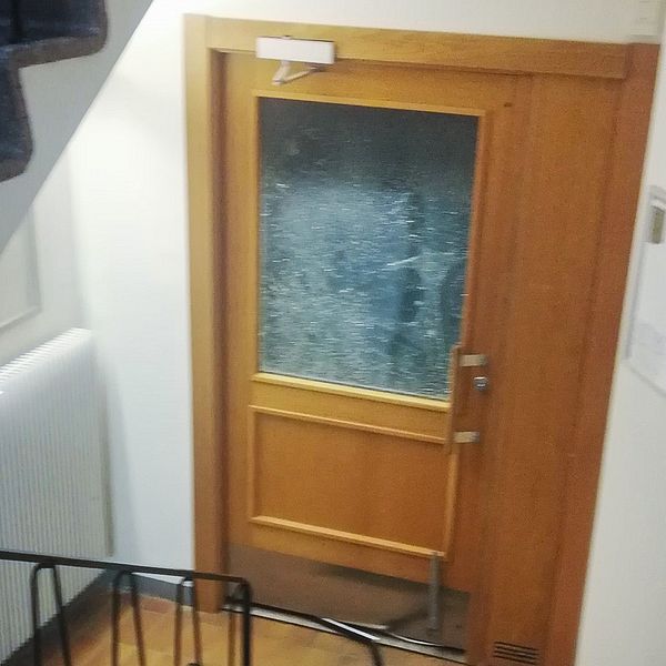 Trasig glasdörr i en trappa, med sprickor och krosskador synliga i glaset, omgiven av en vit vägg och ett element.