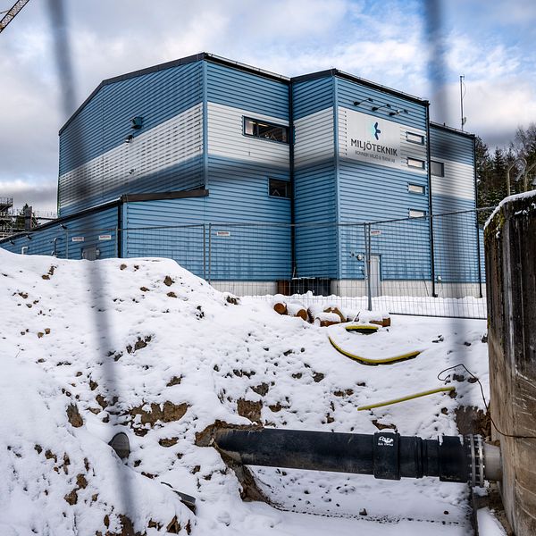 Snötäckt byggarbetsplats med ett blått och silverfärgat industribyggnad, märkt med ”PilotTremin”, synlig i bakgrunden. En stor kran reser sig till vänster och en svart stor rörledning sträcker sig över snön, omgiven av byggstängsel.