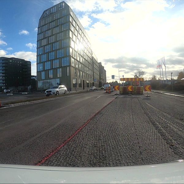 En vägscen som visar pågående vägbygge. I förgrunden syns avstängningskoner och byggutrustning, inklusive en grön maskin. Bakgrunden har en modern byggnad och några mindre byggnader. Himlen är delvis molnig med solstrålar som syns.