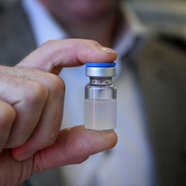 En person håller upp en liten glasampull med en blå kork, innehållande en oklar vätska, mot kameran. Bakgrunden är suddig och antyder en medicinsk miljö.