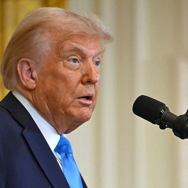Donald Trump står vid en mikrofon i ett inomhusmiljö, med guldfärgad bakgrund.