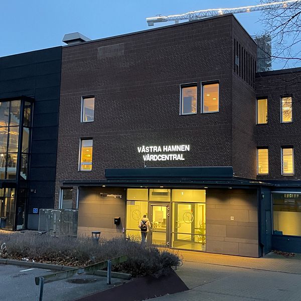 Byggnaden avbildar Västra Hamnen Vårdcentral, som har en modern design med stora fönster som lyser upp fasaden i skymningen. Entrén är välkomnande med en glasdörr och breda steg som leder in. Området är omgivet av grönområden och närliggande byggnader.