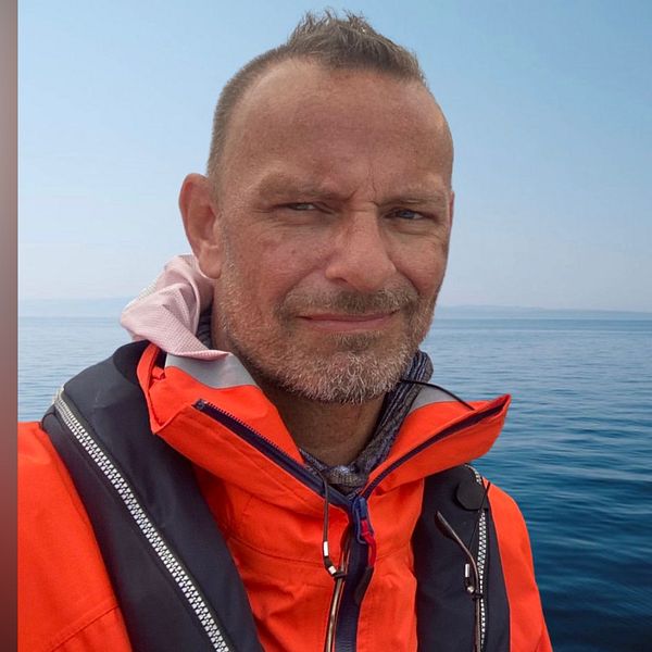 En person i en ljus orange flytväst står framför stilla hav, med en klar himmel i bakgrunden.