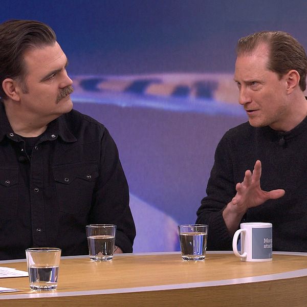 Två personer sitter vid ett runt bord i en TV-studio under ett samtal. Den ena personen gestikulerar med handen, medan den andra lyssnar uppmärksamt. På bordet finns flera glas med vatten och en mugg. En abstrakt, blå bakgrund syns bakom dem som skapar en professionell atmosfär.