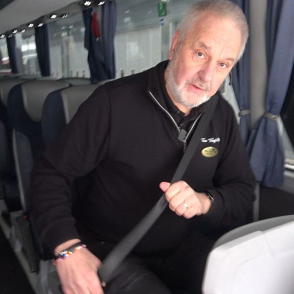 Busschauffören Stefan Wahlén sitter i en buss och fäster säkerhetsbältet, omgiven av tomma säten. Bussens interiör är modern med grå och blå klädslar och belysning i taket.