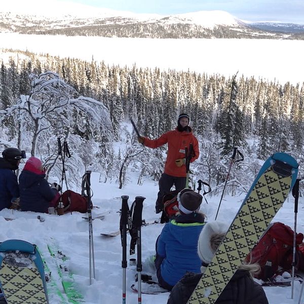 En grupp skidåkare sitter i snön och lyssnar på en guide som pratar framför dem, med en snöklädd skog och berg i bakgrunden. Vintrig landskapssceneri med skidutrustning synlig omkring dem.