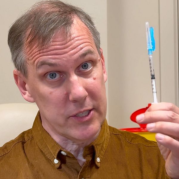 Vetenskapsreporter Johan Bergendorff håller en spruta med blått lock i en klinisk miljö.
