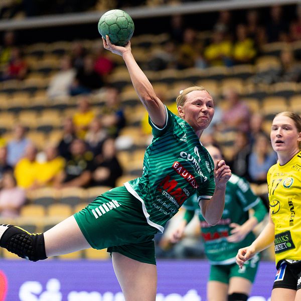 En handbollsspelare i grön tröja och svart shorts kastar bollen mot mål under en match, medan en motståndare i gul tröja observerar från sidan. Publiken i bakgrunden är delvis synlig.