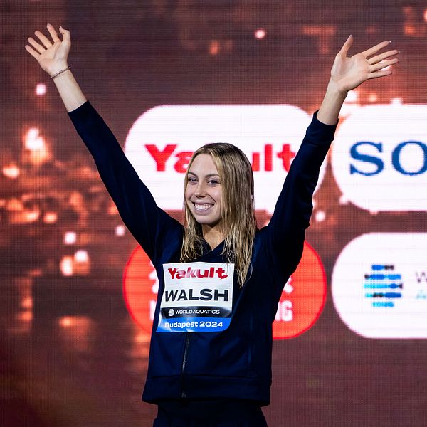 En idrottare sträcker upp sina armar i luften som en gest av seger eller glädje, iförd en mörk outfit med sponsorlogotyper, inklusive ”Yogurt” och ”Walsh.” Bakgrunden visar suddiga ljus och logotyper för företag relaterade till evenemanget.
