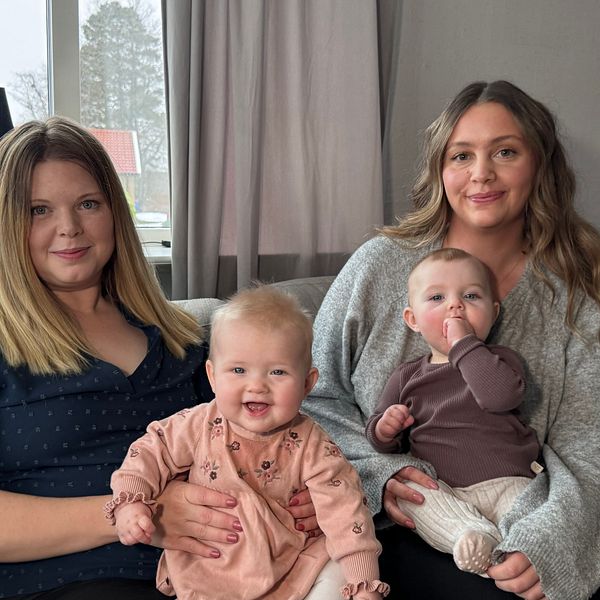 Två kvinnor sitter tillsammans i en vardagsrumsmiljö med fönster i bakgrunden. Den ena kvinnan håller en baby i sitt knä, medan den andra kvinnan också har en baby sittande i knät. Rummet har mjuka färger och ljus inredning som ger en hemtrevlig känsla.