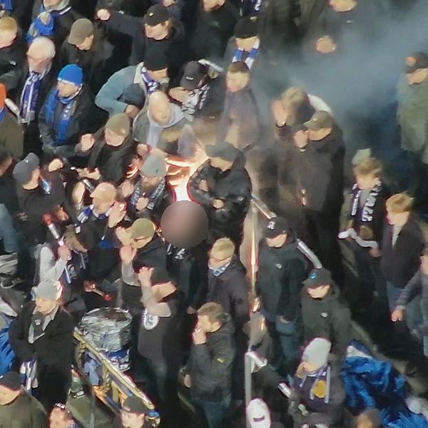 Folksamling klädd i mörka kläder, med någon hållande en fackla, omgiven av rökmoln.