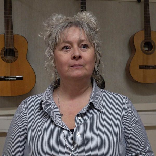 En kvinna sitter och ser sammanbiten ut, bakom hänger flera gitarrer.