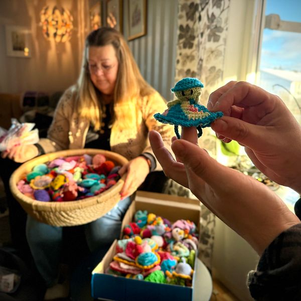 En person håller upp en färgglad virkade figur med en grön och blå klänning. I bakgrunden sitter en annan person med en stor korg fylld med fler virkade figurer i olika färger och former. Rummet är ljust med fönster mot en naturlandskap.