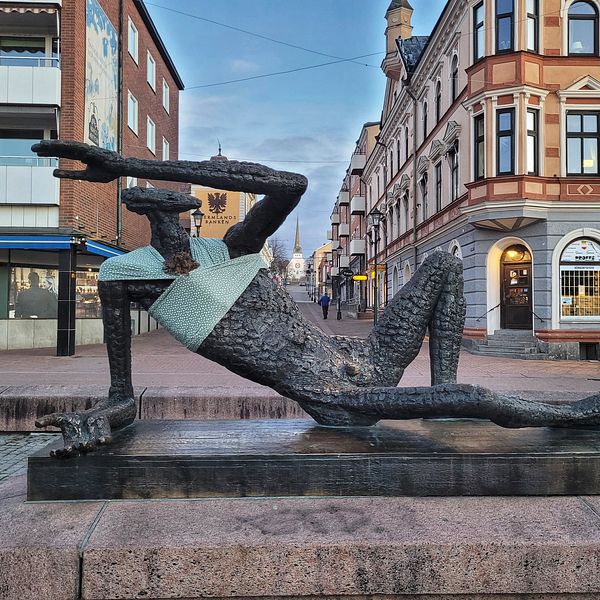 Skulptur av en liggande figur i en stadsmiljö, som pekar åt vänster och bär en ljus tröja. Omgiven av byggnader och gator med affärer i bakgrunden.
