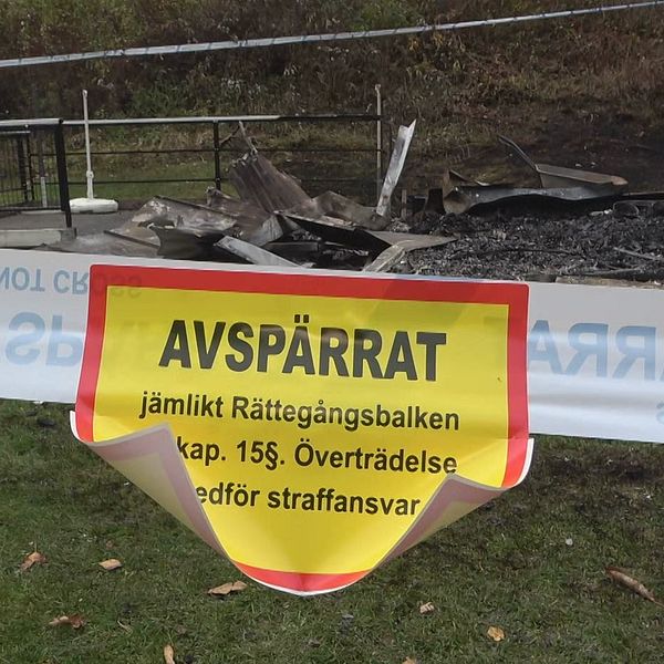 Avspärrat band med en tydlig varningstext som informerar om att området är stängt enligt Rättegångsbalken, i bakgrunden syns förbrända rester av möbler och en askaområde.