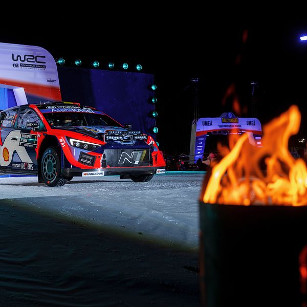 En rallybil deltar i ett nattligt event under Rally Sweden, med ett eldsken i förgrunden som skapar dramatisk belysning. Bakgrunden visar en belyst scen med skyltar och ytterligare en rallybil.