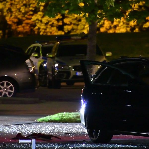 Nattbild av en upplyst parkering där en svart bil med öppen dörr syns i förgrunden. Glassplitter ligger på marken i närheten av bilen. I bakgrunden syns fler parkerade bilar och ett träd med belysning.