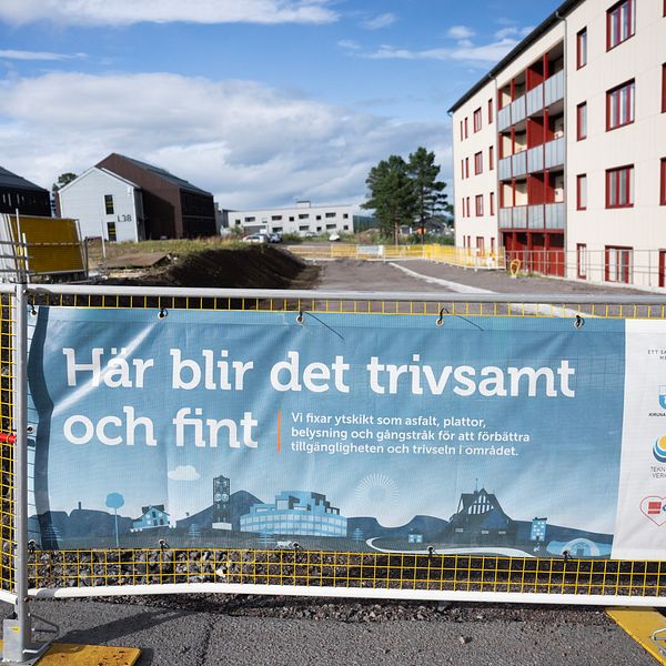 Byggarbetsplats med skylt om trivsam nyproduktion och bostadshus i bakgrunden.