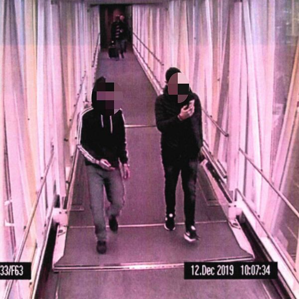 Två män går igenom en korridor på en flygplats, fotograferade av en bevakningskamera.