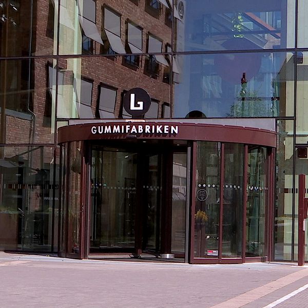 Entrén till Gummifabriken, en modern byggnad med stora glasytor och en roterande dörr, omgiven av stadsmiljö och blomsterarrangemang.