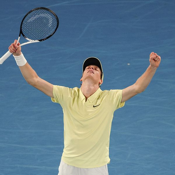 En tennisspelare iklädd ljusgul tröja och vit keps firar en seger på tennisbanan med uppsträckta armar, medan han håller i ett tennisracket.