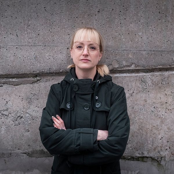 En person står med armarna i kors framför en sliten och fläckad stenvägg. Personen bär en mörk jacka och har sitt ljusa hår uppsatt i en knut.