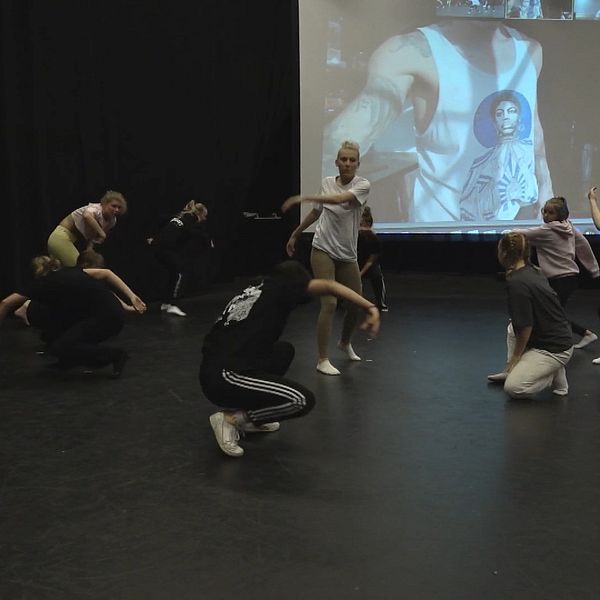 Grupp av dansare från sommardrömjobb projektet som övar på en scen, med en stor projektor i bakgrunden som visar en bild av en person. Dansarna utför olika rörelser, några sitter på golvet medan andra står och rör sig. Lokalen har mörka väggar och en enkel, svart golvbeläggning.