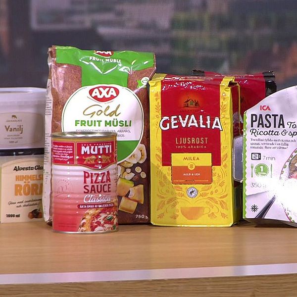 Produkter från olika livsmedelsmärken står uppställda på ett bord. Från vänster till höger syns en burk med vaniljkräm, en förpackning med frukostmusli från AXA, en burk pizzasås från Mutti, en förpackning kaffe från Gevalia och en färdigpastarätt. Bakgrunden består av en suddig stadsbild.