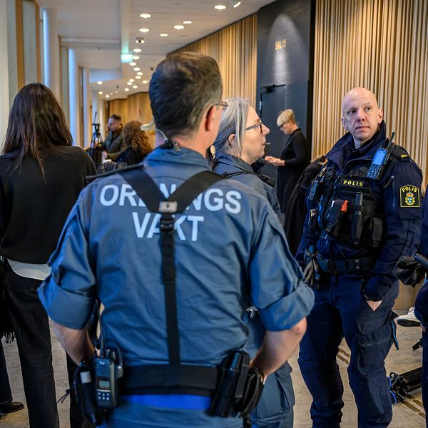 Poliser och bevakningspersonal står i en korridor och diskuterar, medan flera personer i civila kläder observerar situationen. Bakgrunden visar en modern inomhusmiljö med träpanel och bänkar.