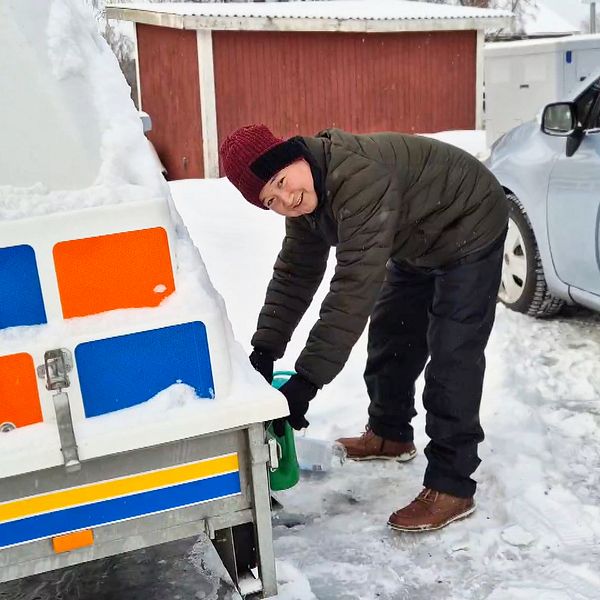 Alidad Zahedy lutar sig över en snötäckt släpvagn och fyller en flaska med vatten från kranen. I bakgrunden syns en parkerad bil och en röd träbyggnad, medan marken är täckt av snö.