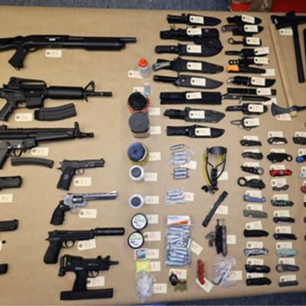 En samling av olika vapen och tillbehör som inkluderar gevär, pistoler, knivar och andra objektspecifikationer, alla märkta med etiketter som anger deras identifiering. Vapen och föremål ligger på en bänkskiva i ordning för att tydliggöra deras typ och egenskaper.