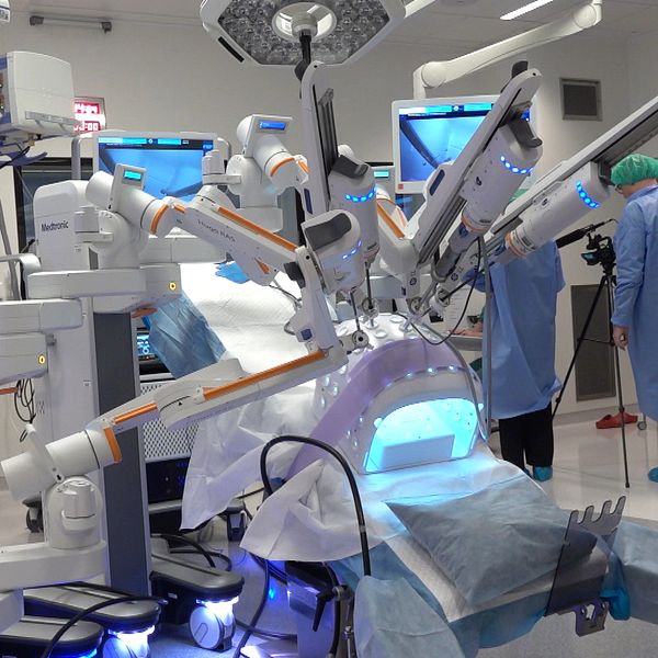 Ett modernt operationsrum med avancerad medicinsk utrustning, inklusive robotkirurgi-maskiner och flera monitorer som visar kirurgiska bilder. I bakgrunden syns medarbetare i kirurgiska kläder och skyddande huvudbonader som förbereder sig för en operation. Belysningen och miljön är sterila och kliniska.