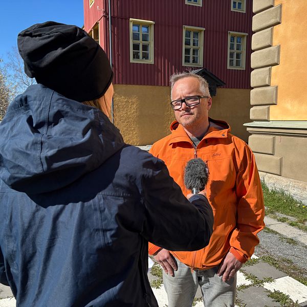 En reporter intervjuar en person utomhus framför färgglada byggnader.