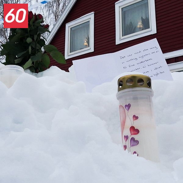 Snö framför en röd träbyggnad med ett tänt gravljus, en bukett rosor och ett handskrivet brev.