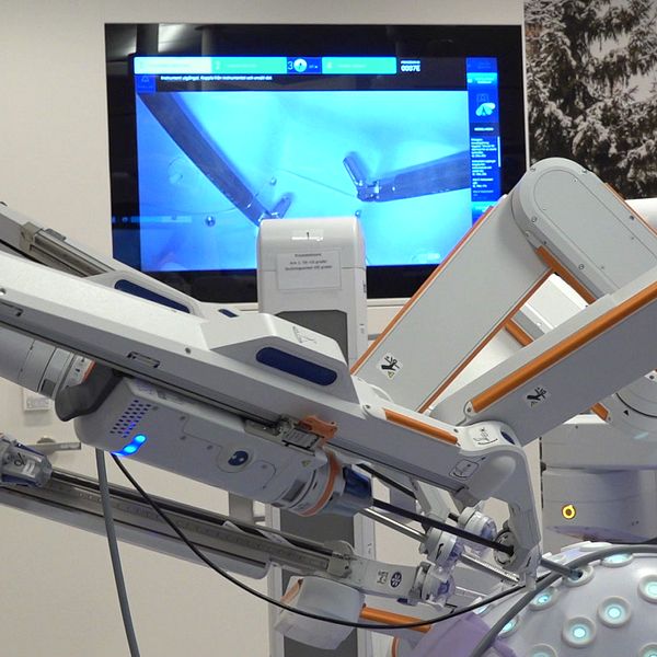 Robotassisterad kirurgiutrustning med flera rörliga delar i förgrunden, och en skärm i bakgrunden som visar en kirurgisk procedur. Utrustningen har blåa lysdioder och är placerad i ett modernt operationsrum.