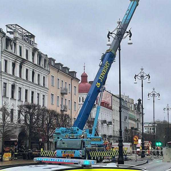 En kran på en stadsgata som används för att lyfta material över byggnader, med flerfärgade byggnader i bakgrunden och en grön och gul räddningsbil i förgrunden. Temperaturen verkar vara kallt och himlen är molnig.