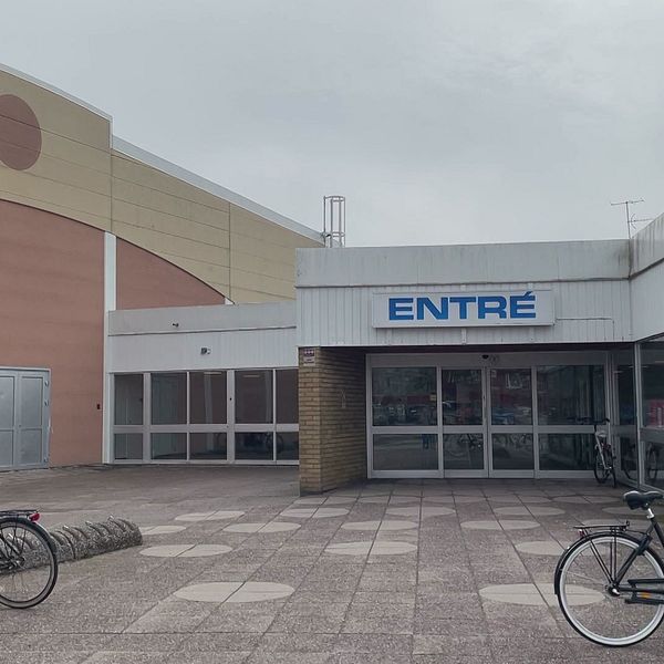 Entrén till en byggnad med glasdörrar, text ”ENTRÉ” ovanför. Två cyklar står parkerade på varsin sida om ingången på en öde stenlagd plats.