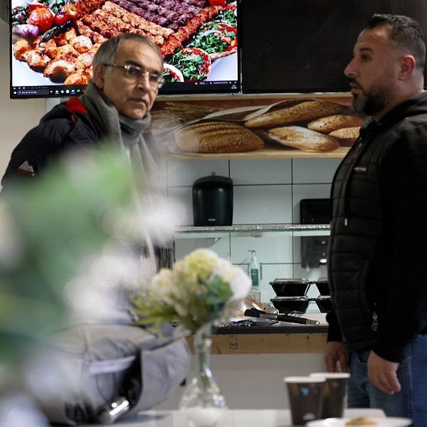 Två personer står och pratar i en restaurangmiljö. I bakgrunden syns en digital meny på skärmar som visar olika grillrätter. En blombukett och kaffemugg står på ett bord i förgrunden.